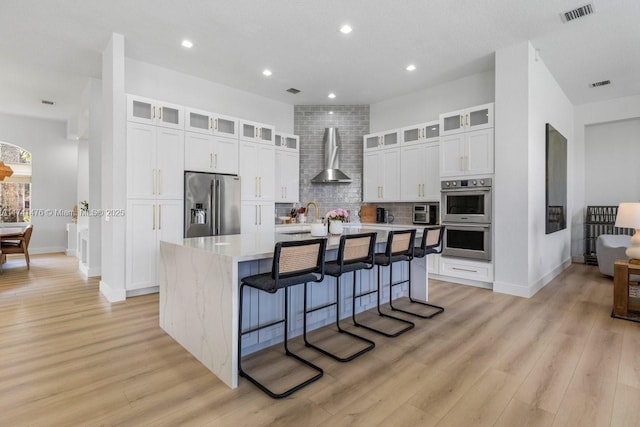 kitchen with appliances with stainless steel finishes, a breakfast bar area, white cabinets, a center island with sink, and wall chimney range hood