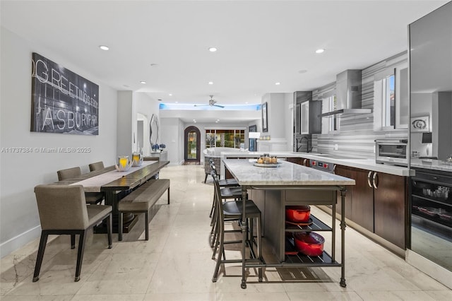 kitchen with dark brown cabinets, a kitchen breakfast bar, light stone countertops, decorative backsplash, and wall chimney range hood