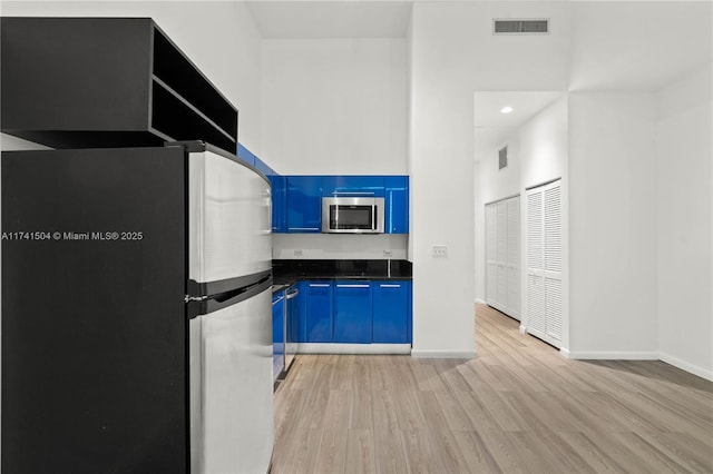 kitchen with stainless steel appliances, blue cabinetry, light wood-type flooring, and a towering ceiling