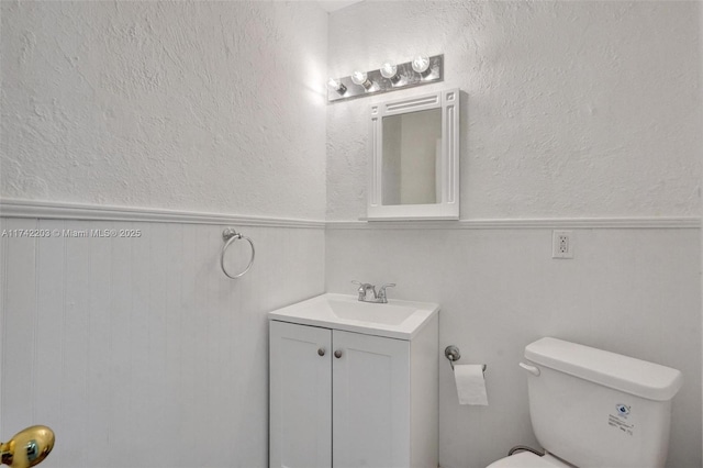 half bathroom featuring a textured wall, toilet, vanity, and a wainscoted wall
