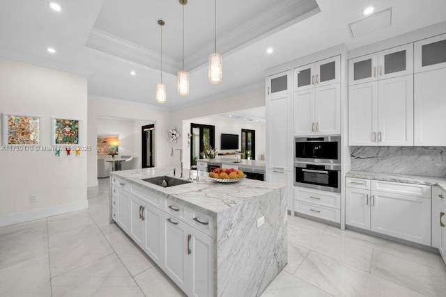 kitchen with sink, light stone counters, a tray ceiling, white cabinets, and a center island with sink