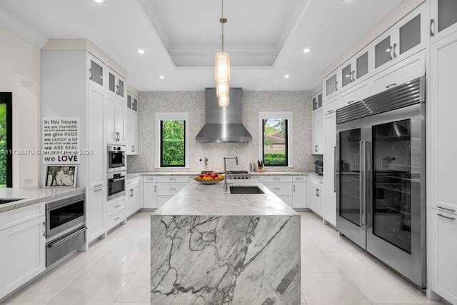 kitchen with wall chimney range hood, a kitchen island with sink, white cabinetry, built in appliances, and a tray ceiling