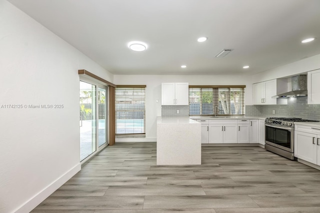 kitchen with wall chimney range hood, backsplash, stainless steel gas range, and white cabinets