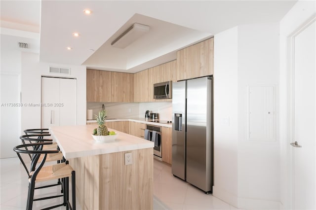 kitchen with light tile patterned floors, stainless steel appliances, a center island, decorative backsplash, and light brown cabinets