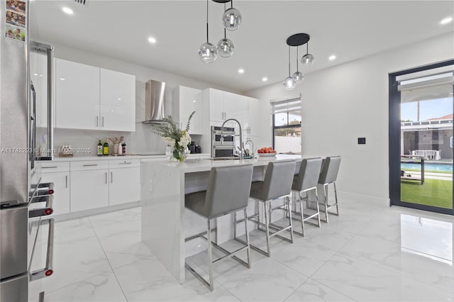 kitchen featuring decorative light fixtures, white cabinetry, an island with sink, plenty of natural light, and wall chimney range hood