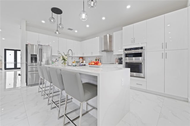 kitchen with decorative light fixtures, wall chimney range hood, an island with sink, stainless steel appliances, and white cabinets