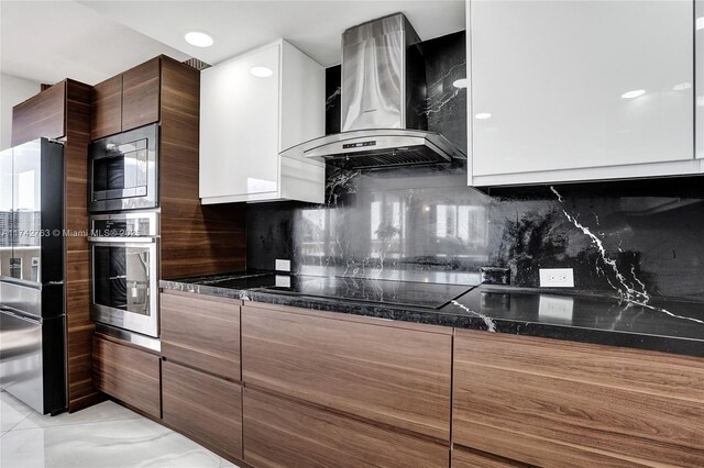 kitchen with wall chimney range hood, white cabinetry, stainless steel appliances, decorative backsplash, and dark stone counters