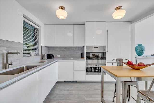 kitchen with double oven, tasteful backsplash, white cabinetry, sink, and light wood-type flooring