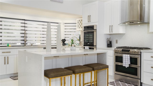 kitchen with wall chimney range hood, appliances with stainless steel finishes, a kitchen breakfast bar, a center island, and white cabinets