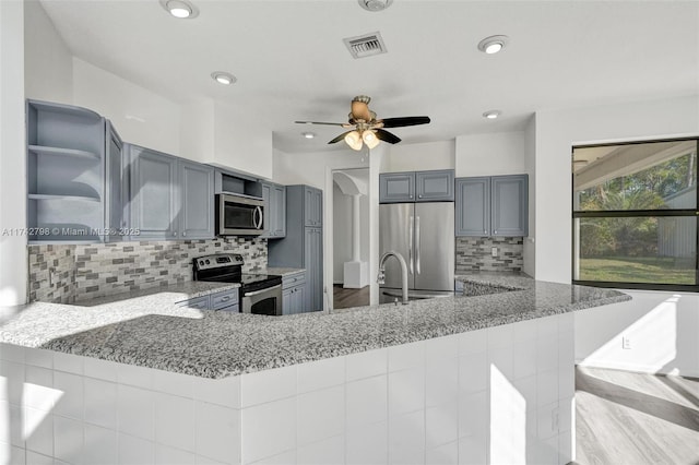 kitchen featuring gray cabinets, decorative backsplash, kitchen peninsula, stainless steel appliances, and light stone countertops