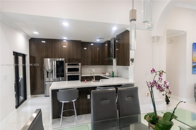kitchen featuring wall chimney range hood, appliances with stainless steel finishes, a kitchen breakfast bar, dark brown cabinetry, and decorative backsplash