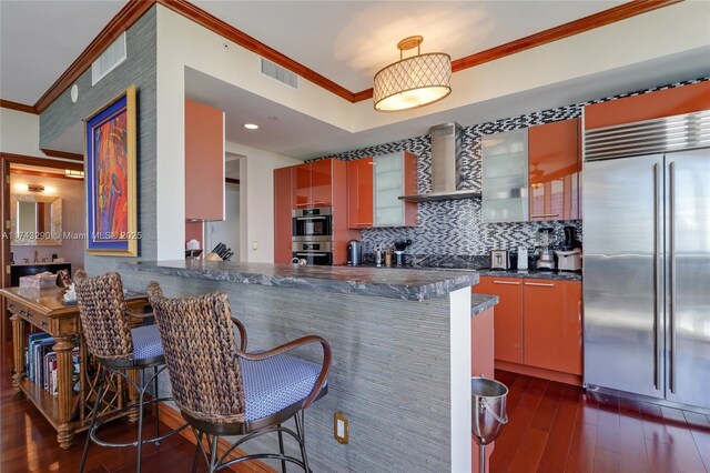 kitchen with ornamental molding, dark wood finished floors, dark countertops, appliances with stainless steel finishes, and wall chimney exhaust hood