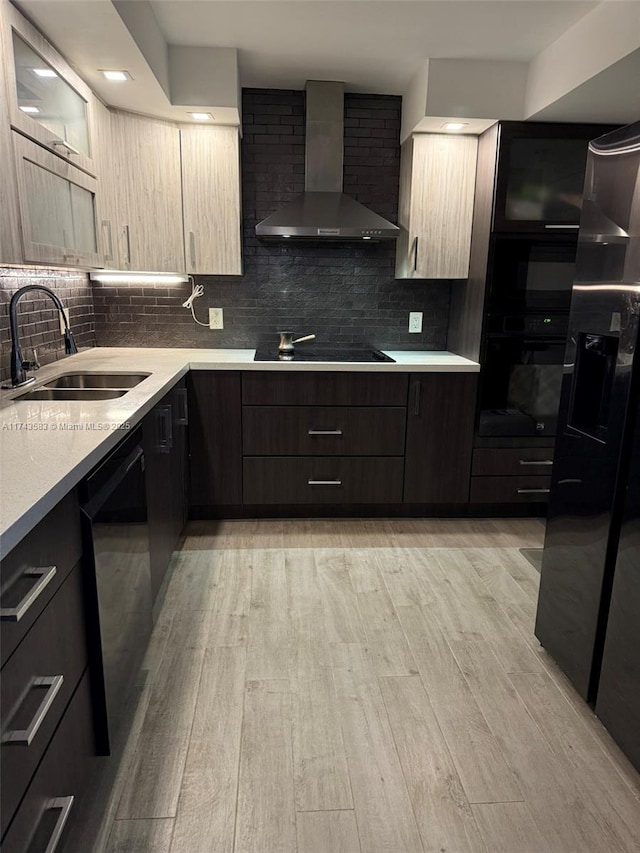 kitchen featuring wall chimney range hood, light countertops, light wood-type flooring, black appliances, and a sink