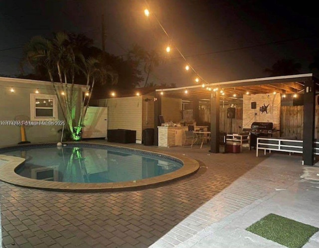 pool at night featuring a grill, a patio area, and an outdoor kitchen