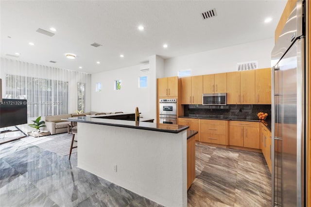 kitchen featuring an AC wall unit, light brown cabinets, an island with sink, stainless steel appliances, and decorative backsplash