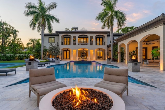 pool at dusk featuring ceiling fan, an outdoor fire pit, and a patio