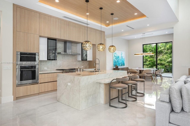 kitchen with sink, decorative light fixtures, a large island with sink, a raised ceiling, and wall chimney range hood