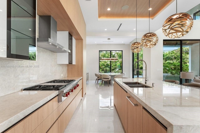 kitchen featuring stainless steel gas stovetop, sink, hanging light fixtures, light stone counters, and wall chimney range hood