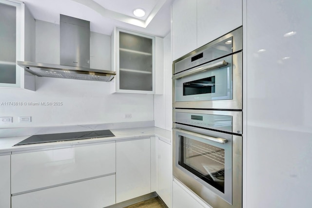 kitchen featuring double oven, white cabinetry, light stone countertops, wall chimney range hood, and black electric cooktop