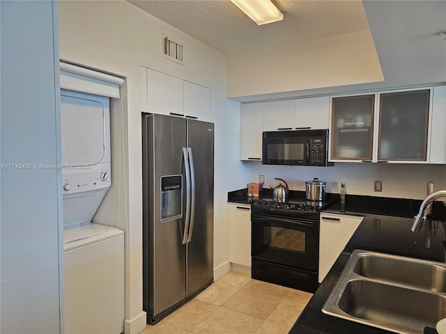 kitchen featuring sink, stacked washing maching and dryer, black appliances, a textured ceiling, and white cabinets