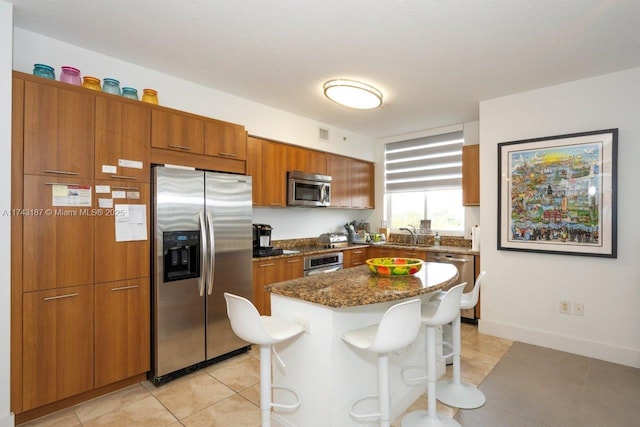 kitchen with dark stone counters, appliances with stainless steel finishes, a breakfast bar area, brown cabinetry, and light tile patterned floors