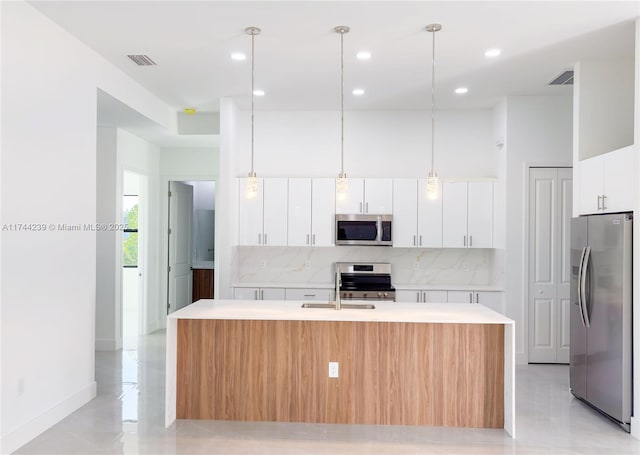 kitchen featuring tasteful backsplash, decorative light fixtures, appliances with stainless steel finishes, a kitchen island with sink, and white cabinets