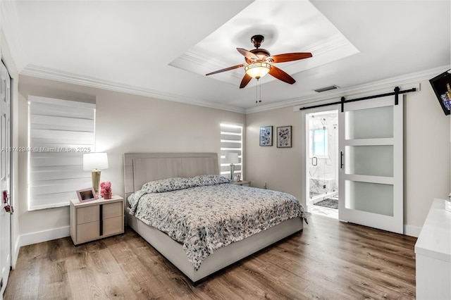 bedroom featuring crown molding, ensuite bath, wood-type flooring, a raised ceiling, and a barn door