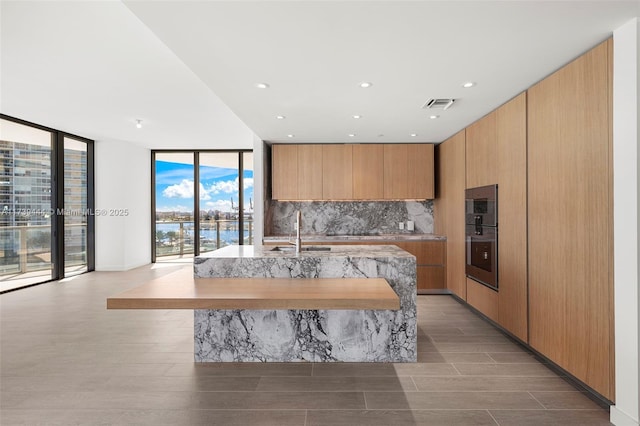 kitchen featuring an island with sink, sink, decorative backsplash, expansive windows, and a water view