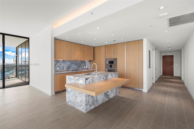 kitchen with sink, a kitchen island with sink, backsplash, dark hardwood / wood-style floors, and light stone countertops