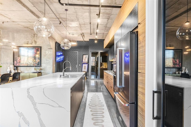 kitchen with light stone countertops, high quality fridge, a center island with sink, and decorative light fixtures