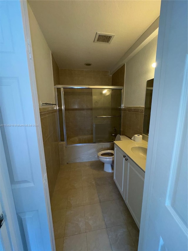 full bathroom featuring toilet, bath / shower combo with glass door, tile walls, vanity, and tile patterned flooring