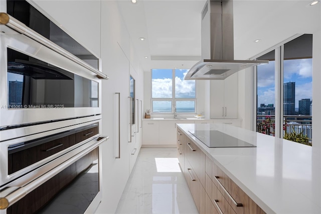 kitchen with light stone counters, island range hood, black electric cooktop, double oven, and white cabinets