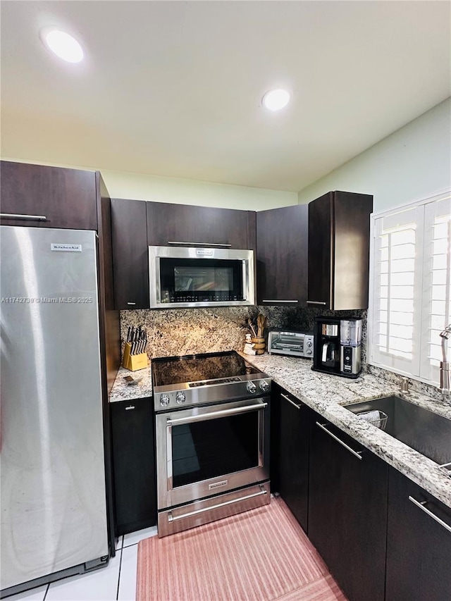 kitchen with appliances with stainless steel finishes, dark brown cabinetry, light stone countertops, light tile patterned floors, and sink