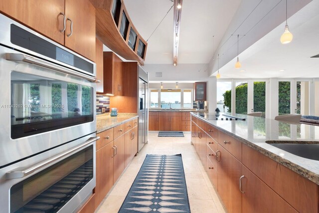 kitchen with brown cabinets, light stone countertops, appliances with stainless steel finishes, light tile patterned floors, and backsplash