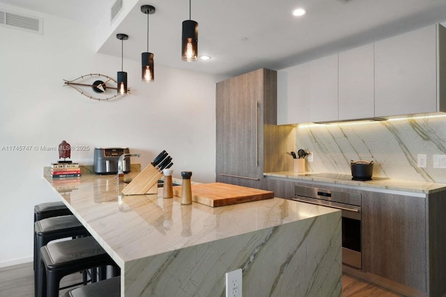 kitchen featuring pendant lighting, backsplash, black electric stovetop, a kitchen bar, and oven
