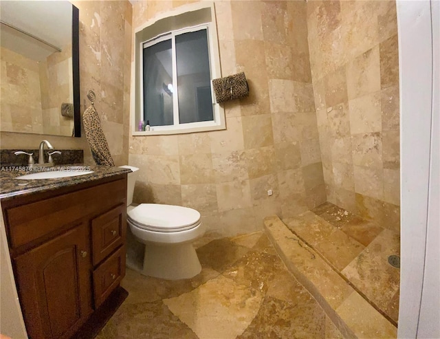bathroom featuring tiled shower, tile walls, toilet, stone finish floor, and vanity