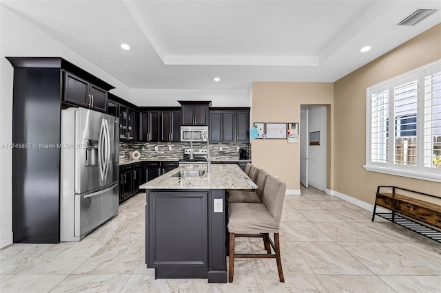 kitchen featuring sink, stainless steel appliances, a tray ceiling, light stone countertops, and a center island with sink