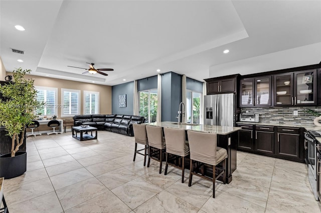 kitchen featuring a breakfast bar, appliances with stainless steel finishes, a raised ceiling, an island with sink, and light stone countertops