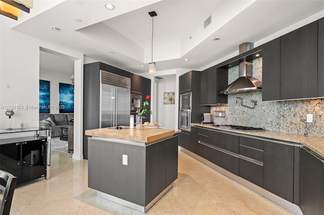 kitchen featuring wall chimney range hood, a kitchen island with sink, built in appliances, decorative light fixtures, and a raised ceiling