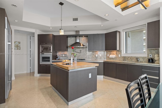 kitchen featuring a raised ceiling, a kitchen island, pendant lighting, stainless steel appliances, and wall chimney range hood