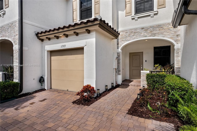 doorway to property featuring a garage, stone siding, a tile roof, decorative driveway, and stucco siding