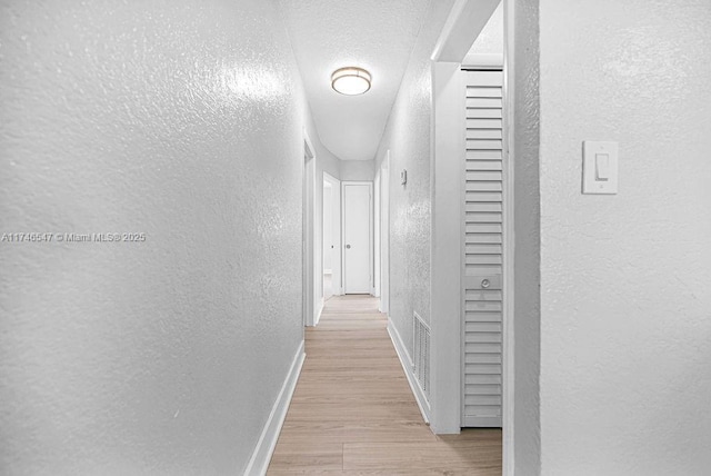 hall with light wood finished floors, visible vents, a textured wall, a textured ceiling, and baseboards