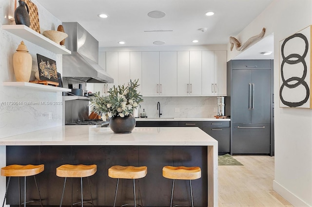 kitchen with built in fridge, a peninsula, wall chimney range hood, open shelves, and a kitchen bar