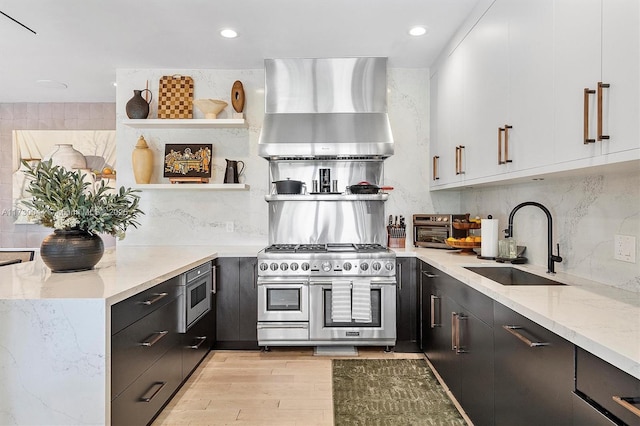 kitchen with wall chimney exhaust hood, appliances with stainless steel finishes, light stone countertops, open shelves, and a sink