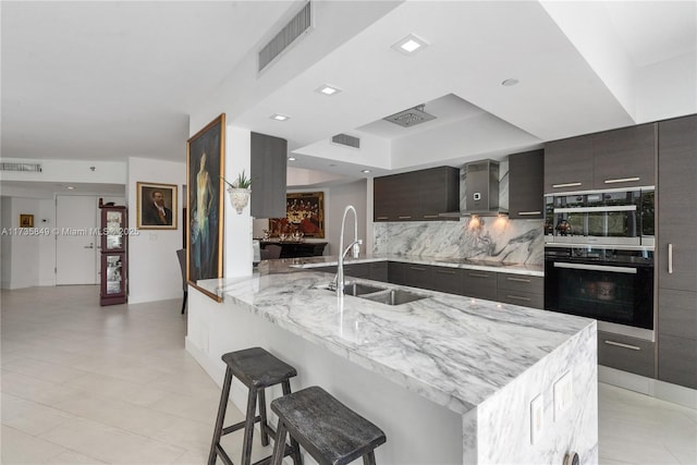 kitchen with sink, a breakfast bar area, double oven, wall chimney range hood, and backsplash