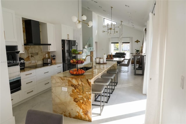 kitchen with tasteful backsplash, a breakfast bar area, wall chimney range hood, black appliances, and white cabinetry