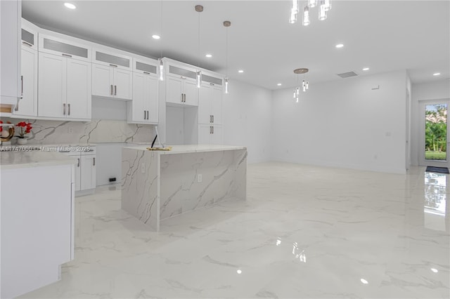 kitchen with tasteful backsplash, hanging light fixtures, a center island with sink, light stone countertops, and white cabinets