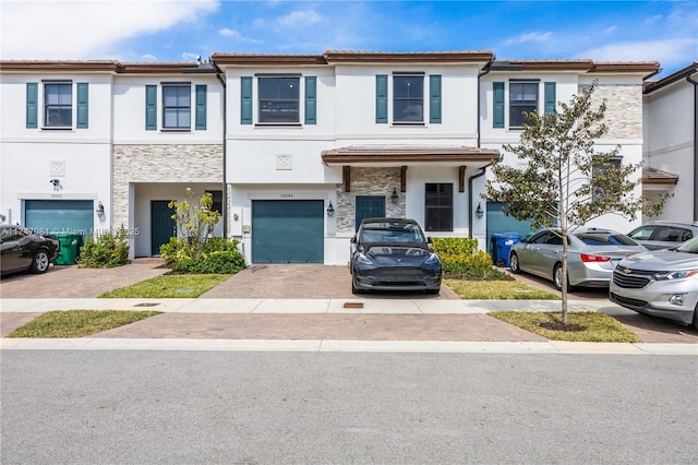 townhome / multi-family property with a garage, stone siding, a tiled roof, and stucco siding