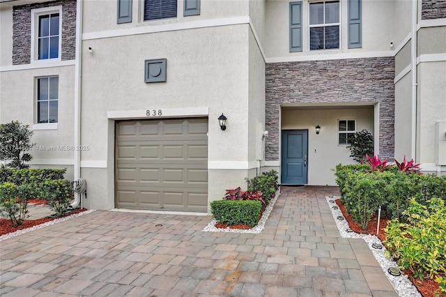 doorway to property featuring an attached garage, stone siding, decorative driveway, and stucco siding