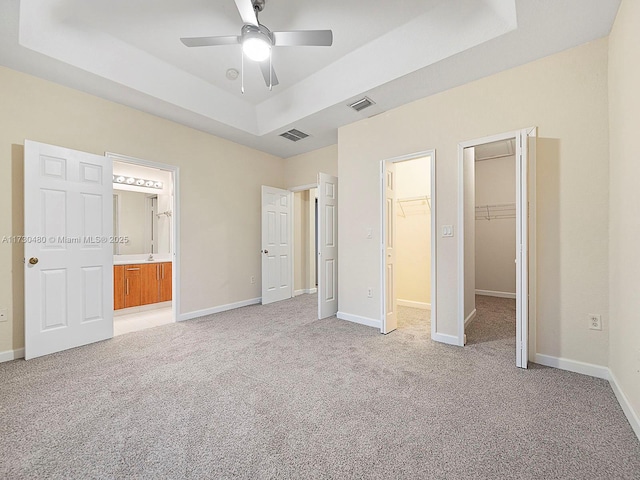 unfurnished bedroom featuring a walk in closet, ensuite bathroom, ceiling fan, a raised ceiling, and light carpet